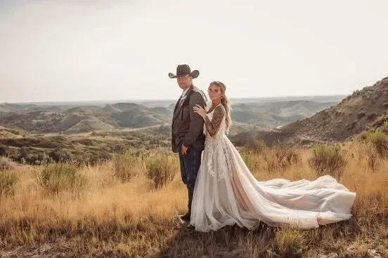 Couple in wedding attire standing in a grassy desert landscape, with a flowing train and rolling hills behind them.