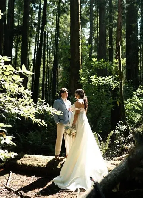 Bride and groom standing together in a sunlit forest, holding hands in wedding attire