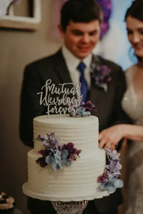 Wedding cake with purple flowers and topper, beside a smiling couple in formal attire