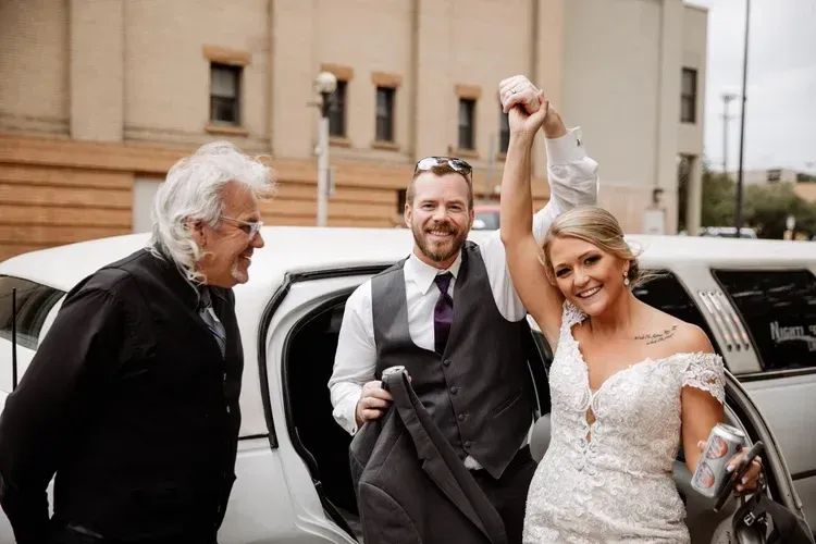 Bride and groom step out of a white limousine, smiling and raising hands in celebration.