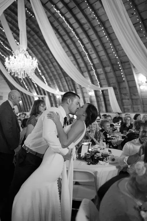 Couple sharing a kiss at a wedding reception under draped fabric and a chandelier, with guests seated nearby