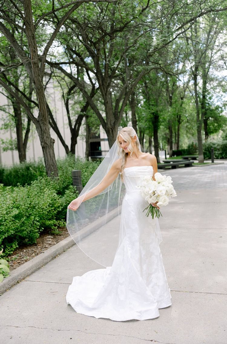 Bride in a strapless white gown holding a bouquet on a tree-lined path