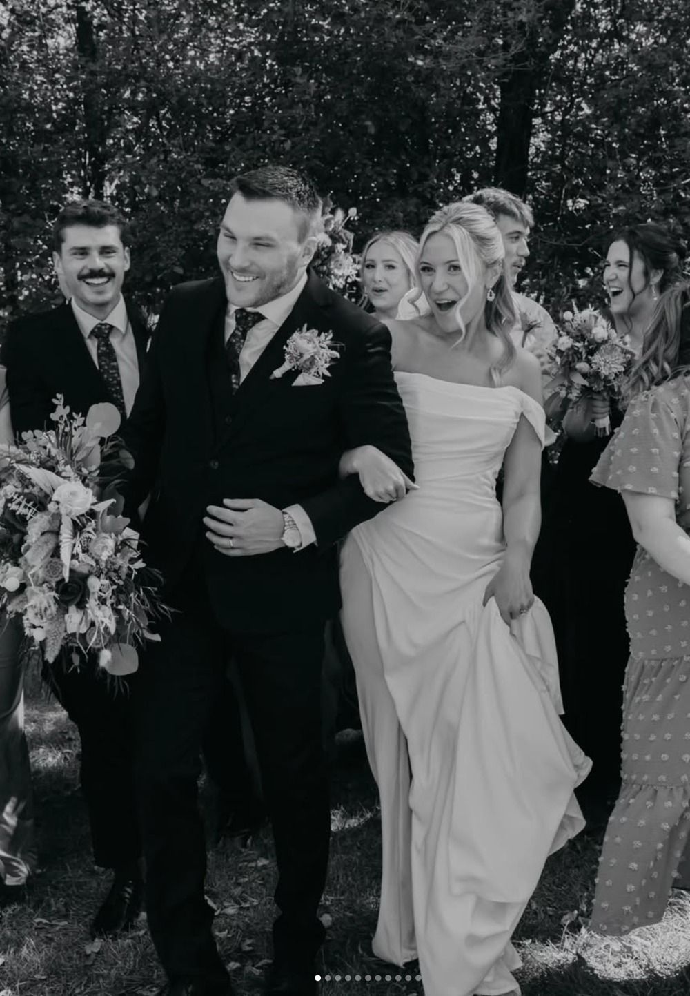 Wedding party walking outdoors, with the bride in an off-shoulder dress holding her gown.