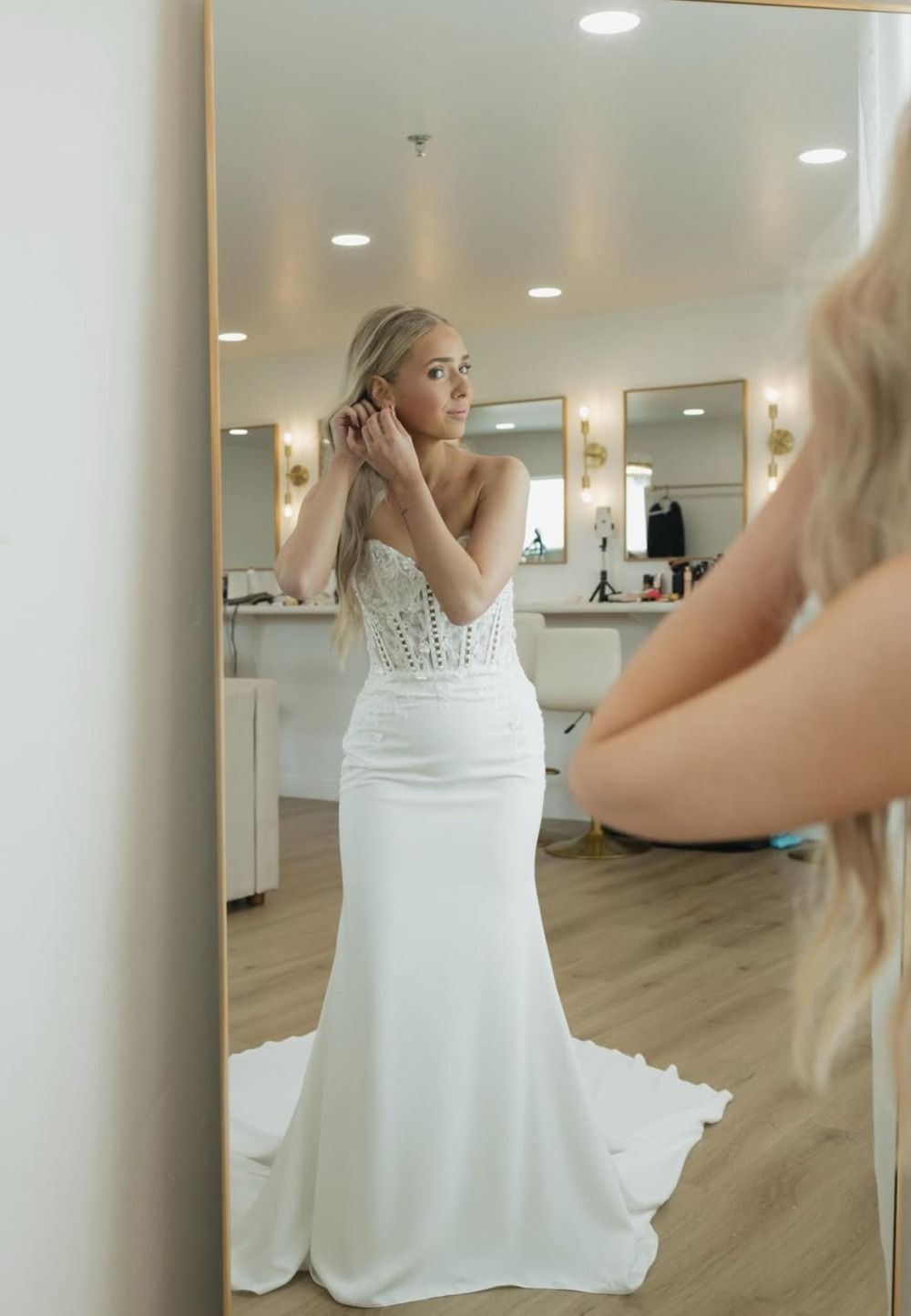 Woman in a white gown adjusting her hair in a mirror inside a bridal shop.