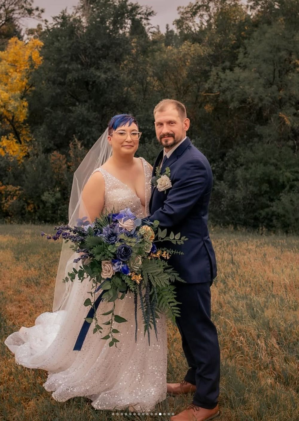 Wedding couple posing outdoors, holding a blue and purple bouquet, with trees in the background