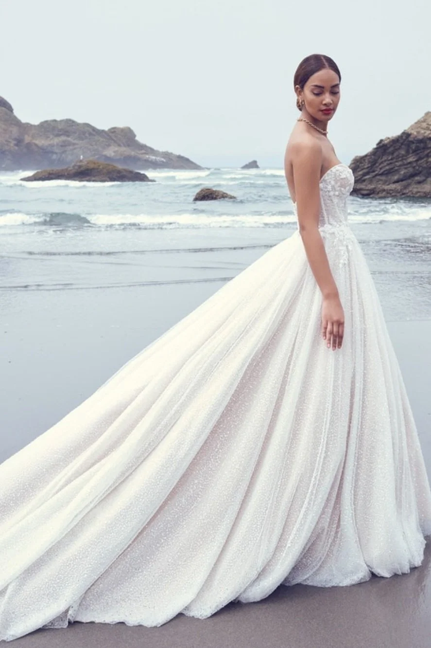 Woman in a white wedding gown standing on a beach with a long flowing train