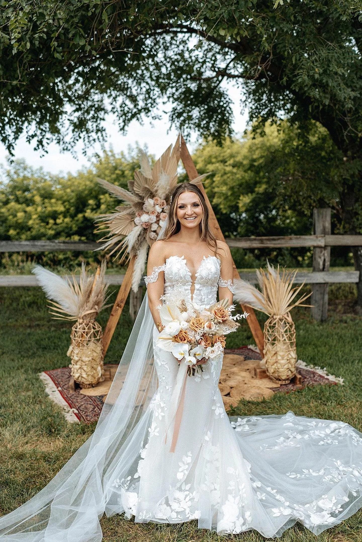 Bride in white gown holding bouquet outdoors under a floral arch with trees and wooden fence behind.