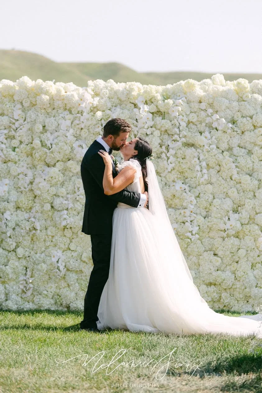 Bride and groom embracing before a white flower wall outdoors