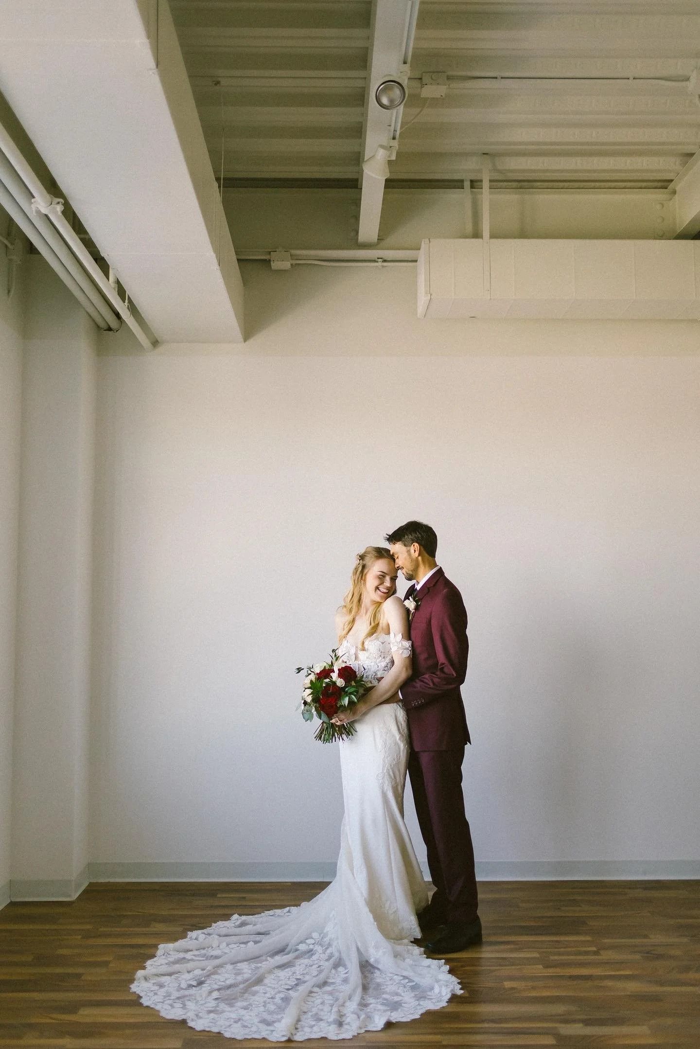 Bride and groom standing together in a bright room, bride holding a bouquet and long train.