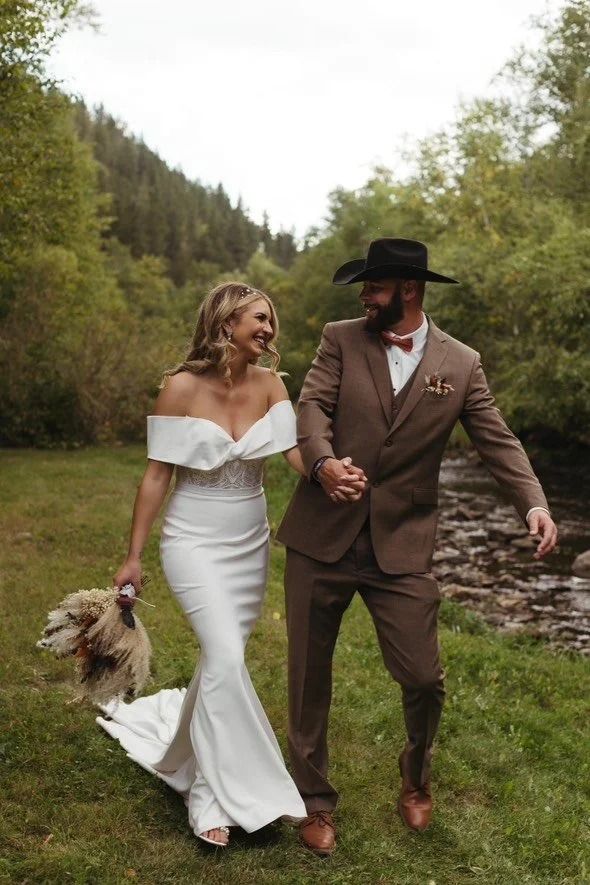 Couple in wedding attire walking hand in hand by a river with a small dog in a grassy forest setting