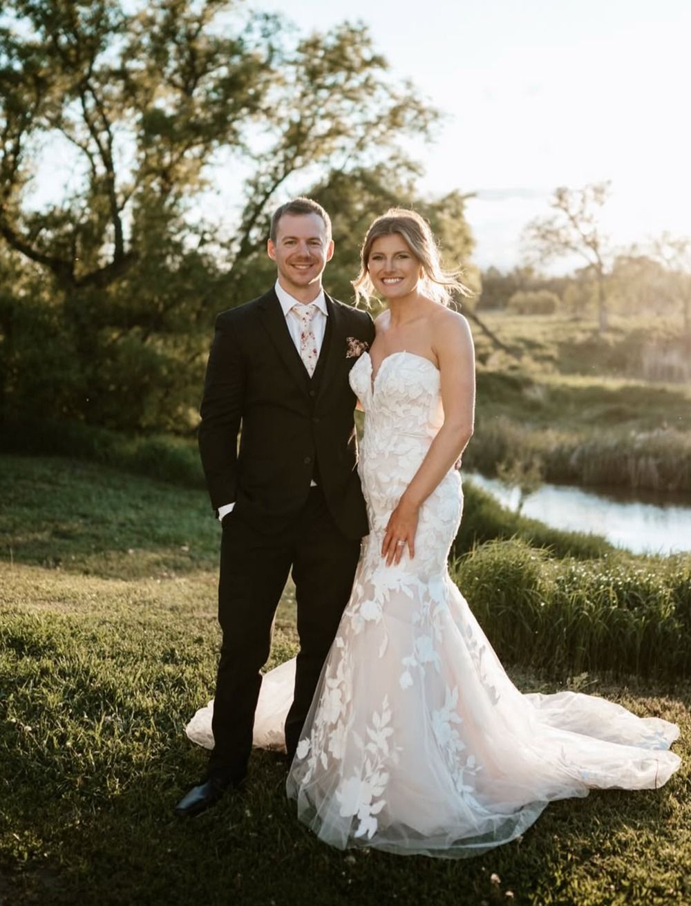 Bride and groom standing outdoors by a pond at sunset, smiling in wedding attire.