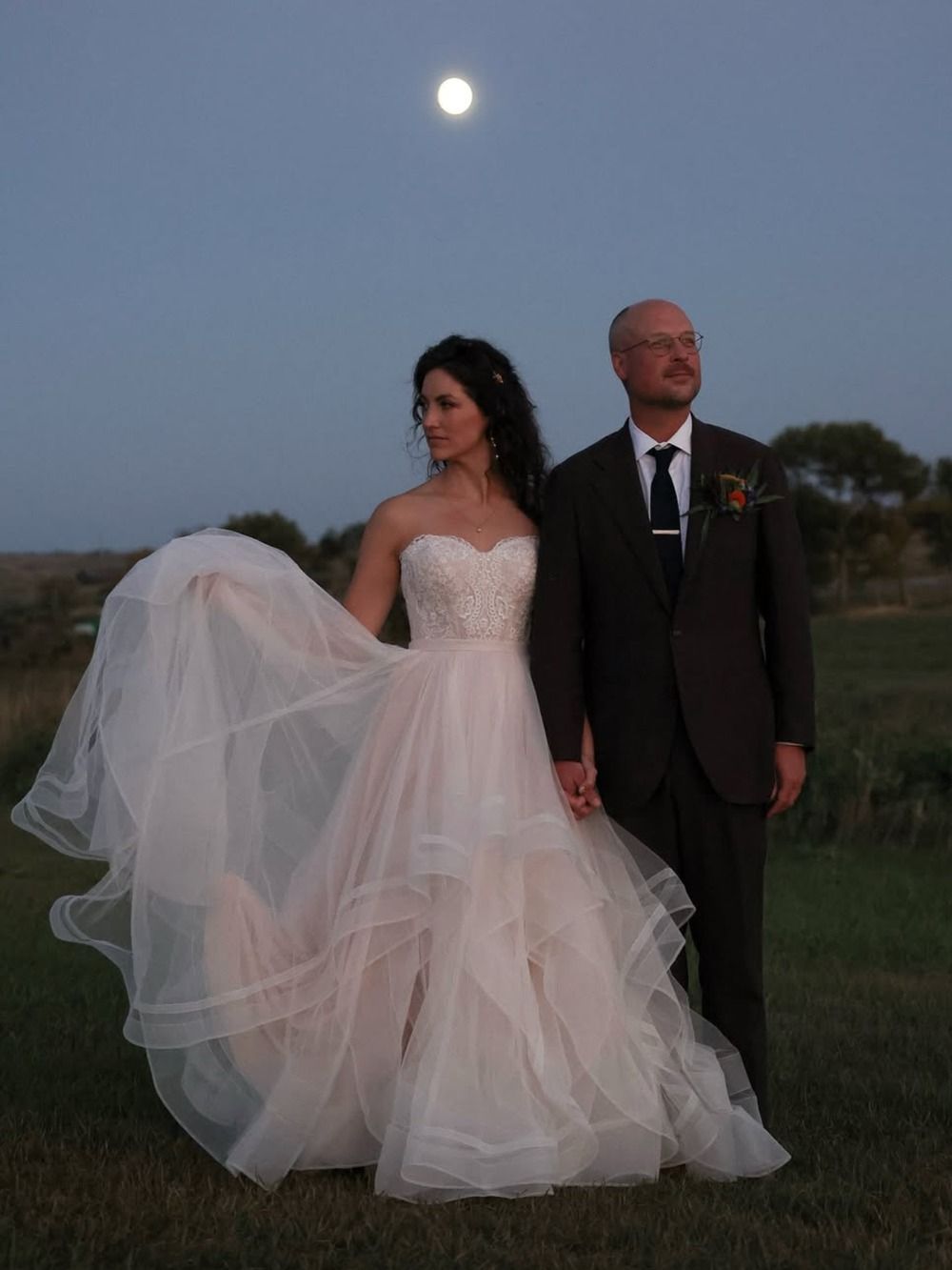 Bride in white gown and groom in dark suit standing outdoors at dusk under a full moon.