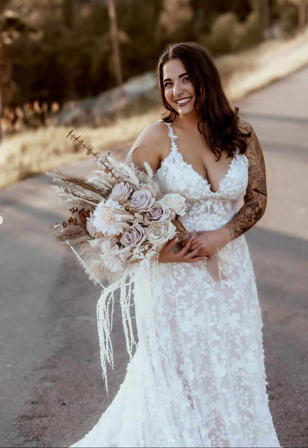 Bride in a white lace gown holding a soft pink bouquet on a road, smiling outdoors.