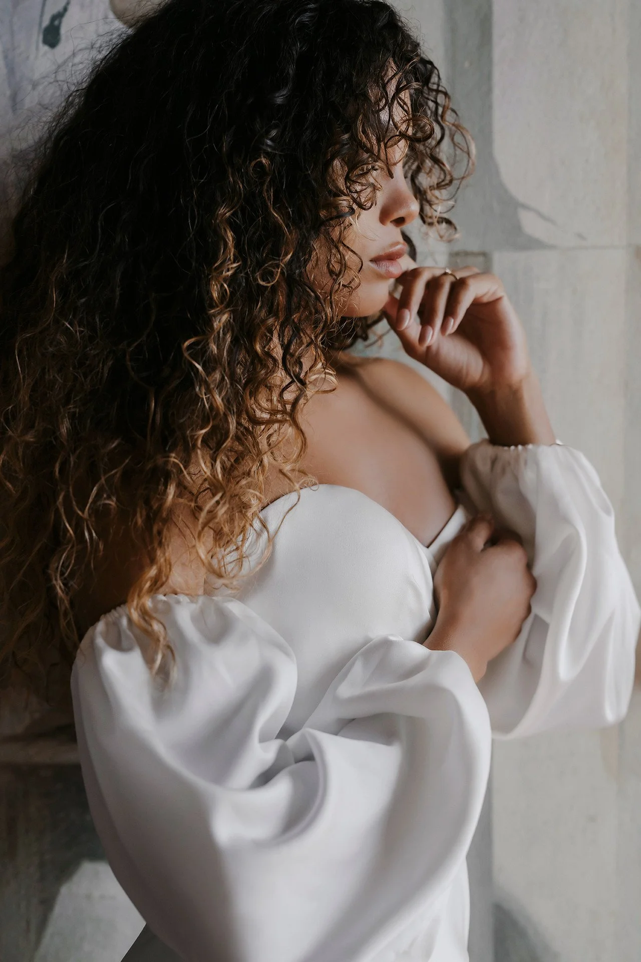 Woman with curly hair in a white off-shoulder blouse, posing by a light wall with hand near her chin