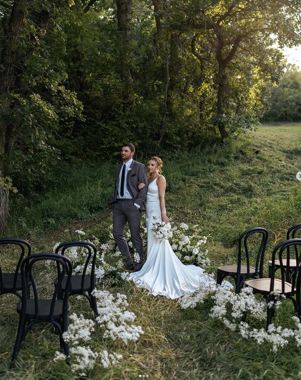 Bride and groom standing in a grassy outdoor aisle with white flowers and black chairs at a woodland wedding