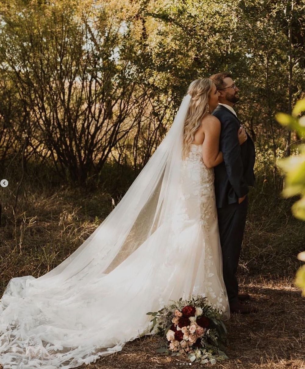 Bride in a long lace gown and groom standing outdoors among trees, with a flowing veil and bouquet nearby