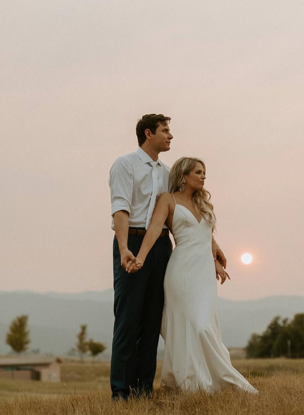Couple holding hands in a field at sunset, with the woman in a white dress and the man in a light shirt.