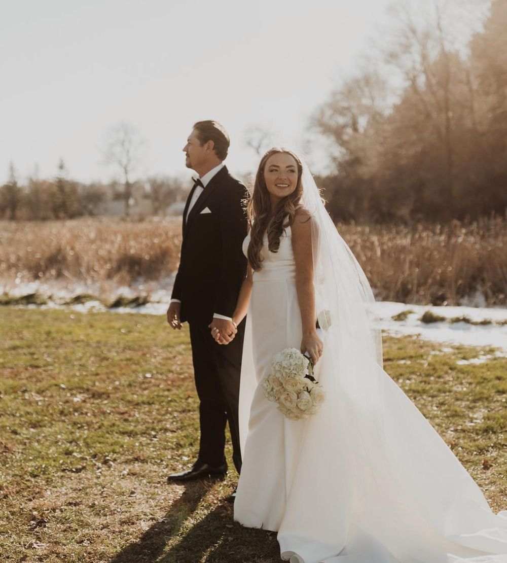Bride and groom holding hands outdoors in a field, with warm autumn trees in the background.