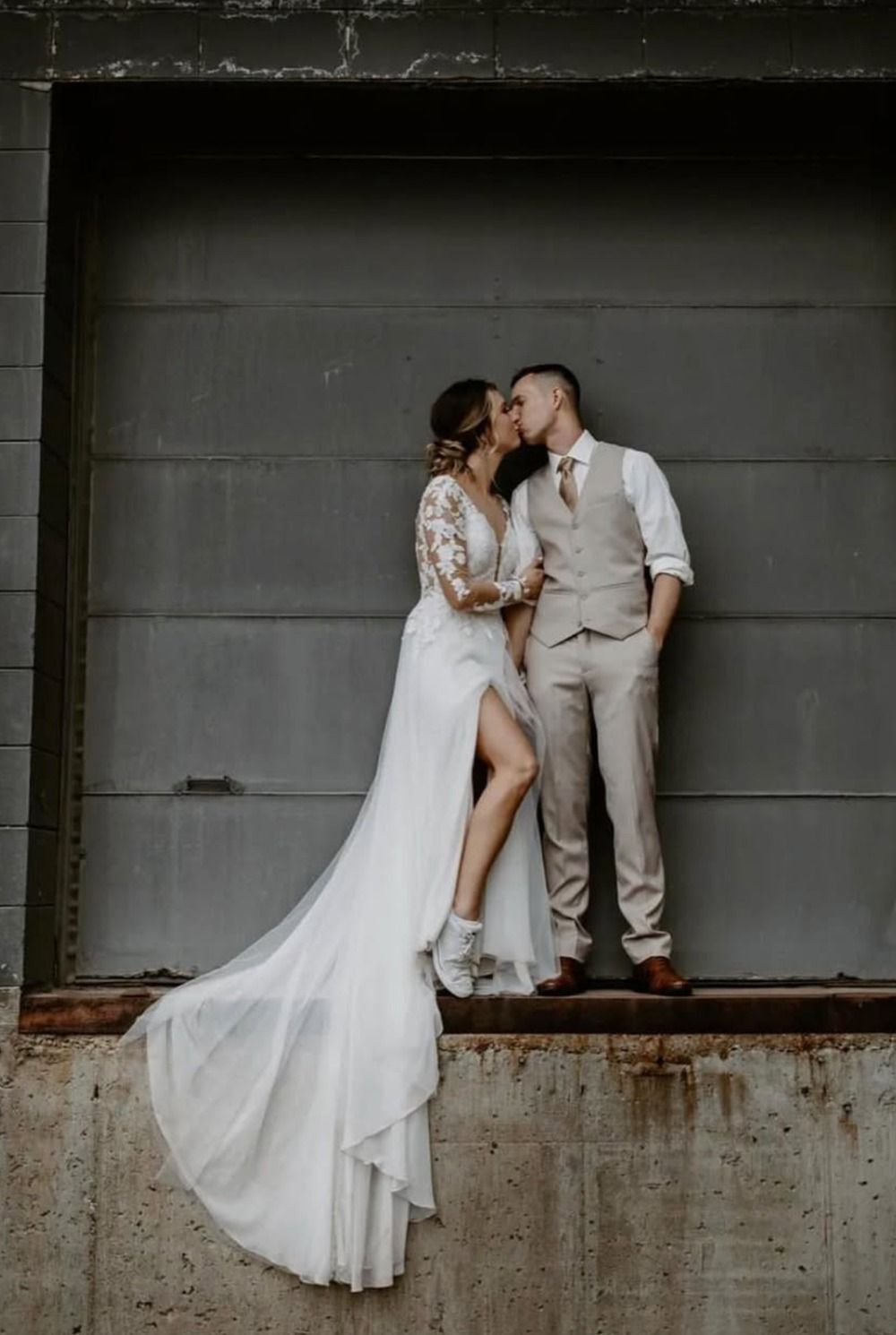 Couple kissing in wedding attire on a concrete ledge against a gray industrial wall