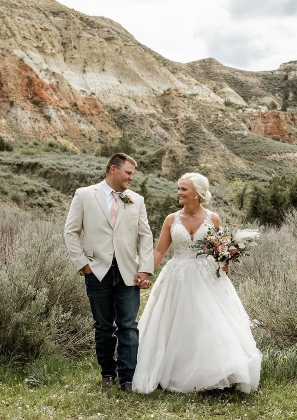 Couple in wedding attire holding hands on a grassy trail beneath rocky hills