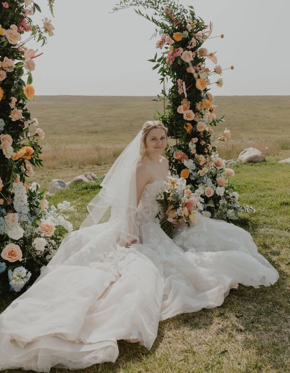 Bride in a white gown sitting beside a flower-covered arch outdoors on a grassy field