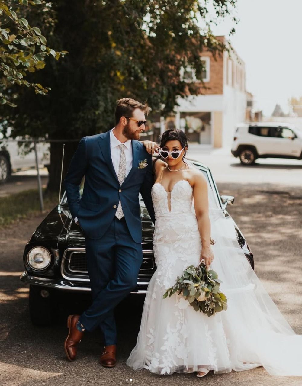 Bride and groom in wedding attire walking beside a vintage car on a street, smiling