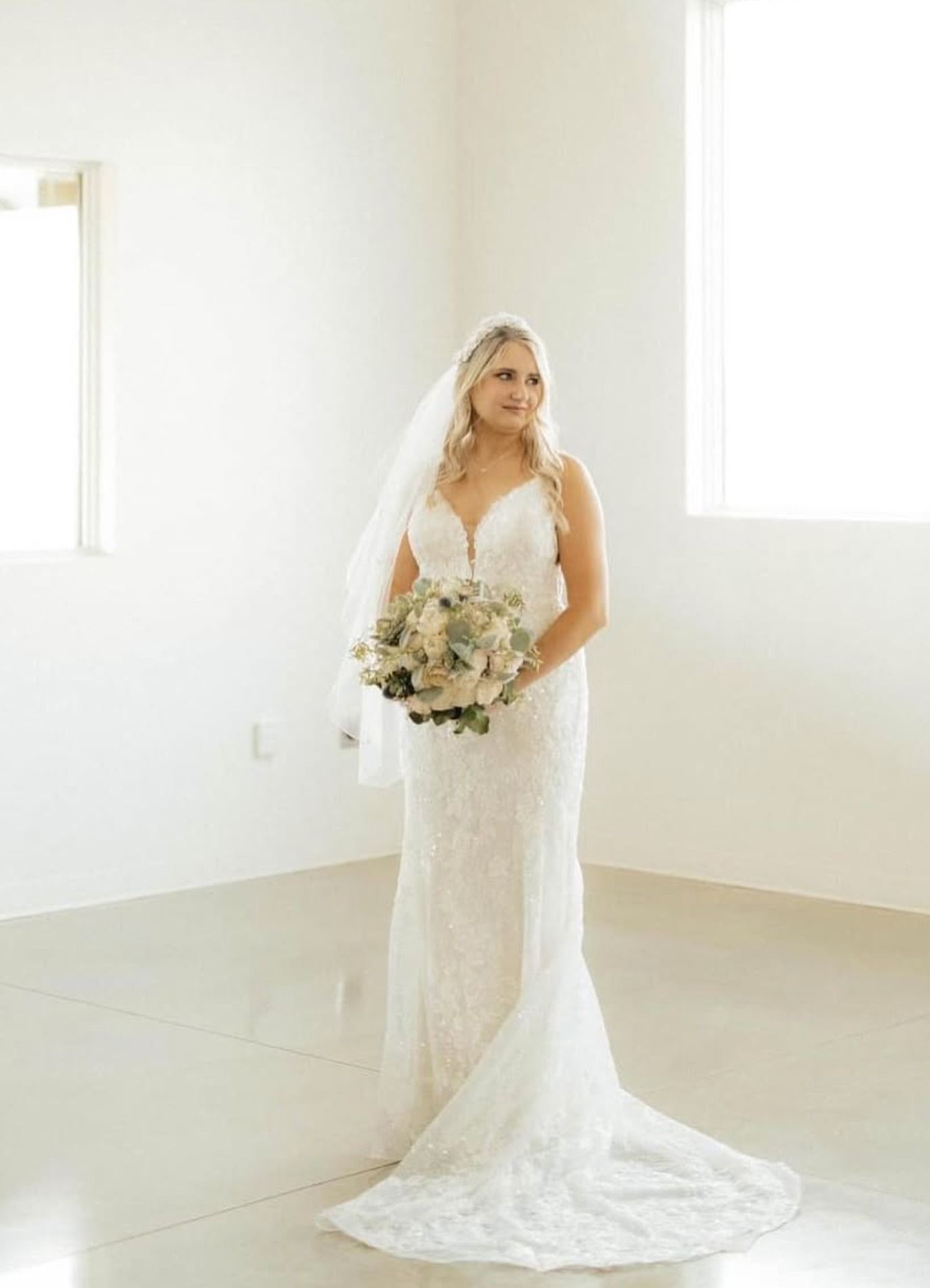 Bride in a white lace wedding dress holding a bouquet in a bright, empty room