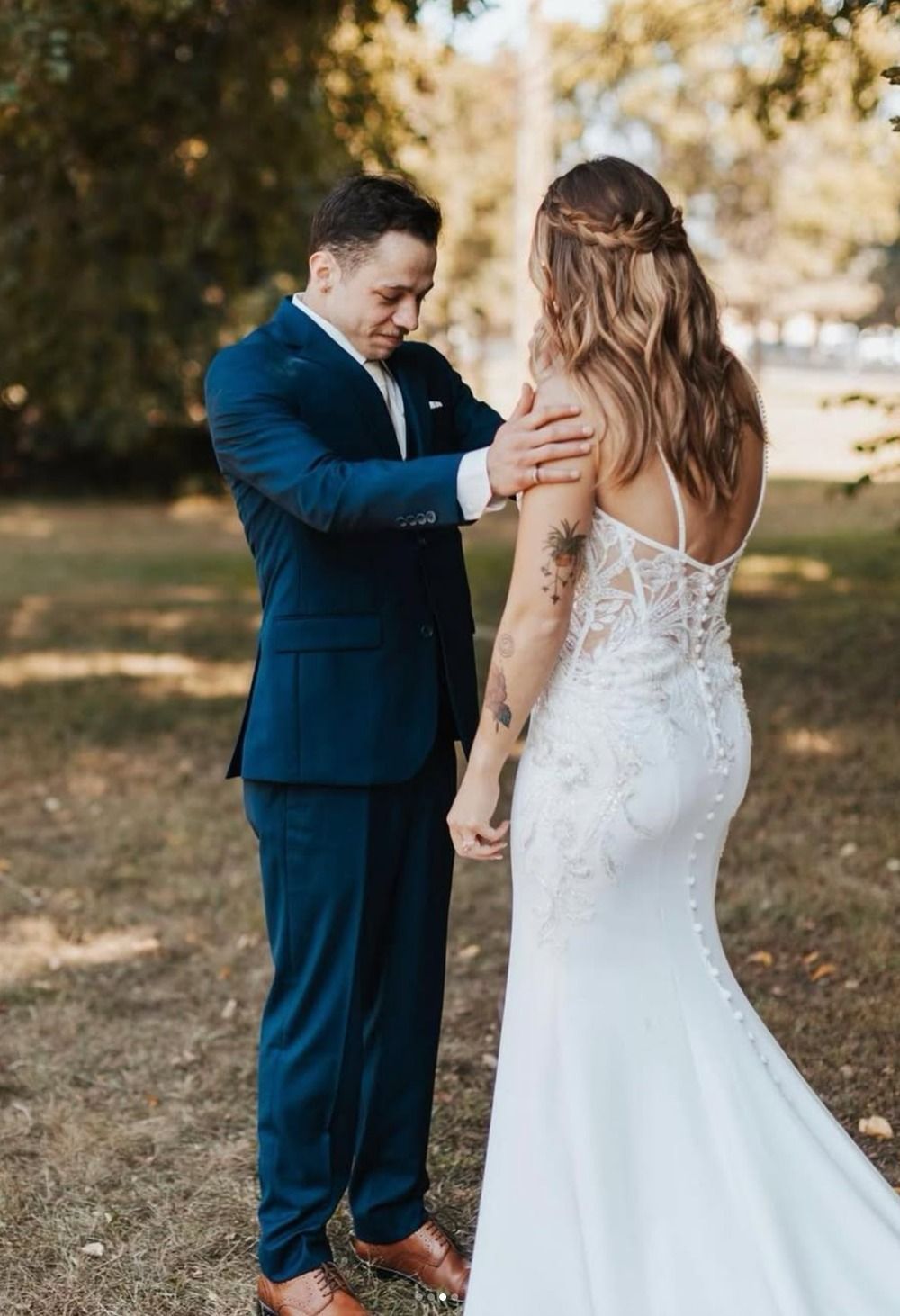 Man adjusting bride’s dress outdoors under trees, with the bride in a white lace gown facing him.
