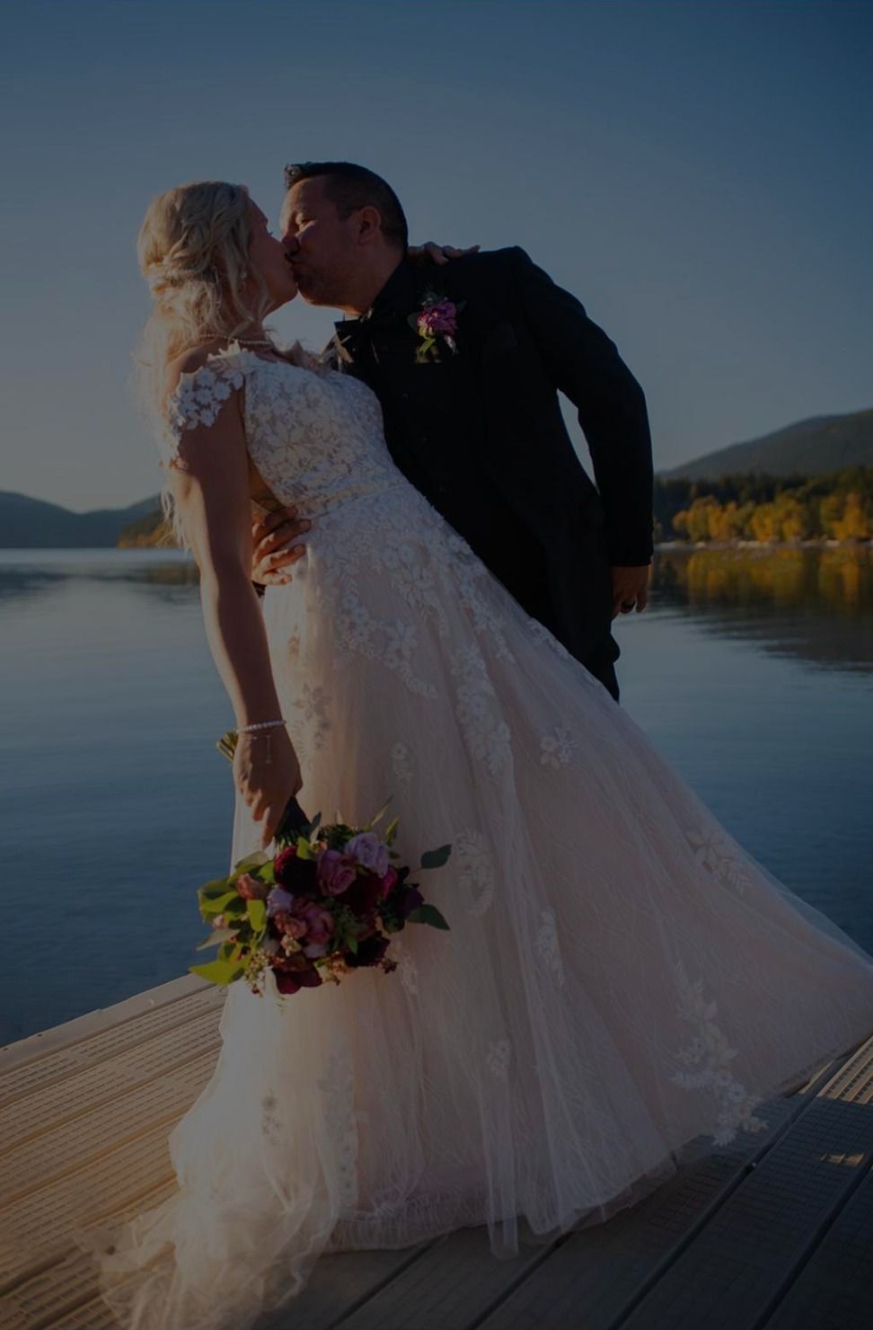 Bride and groom kissing on a lakeside dock at sunset, bride in a white gown holding a bouquet