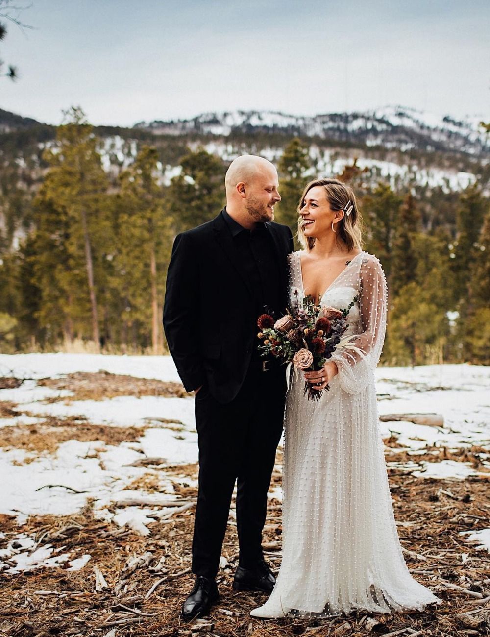 Couple in wedding attire standing together outdoors in a snowy mountain landscape, holding a bouquet.