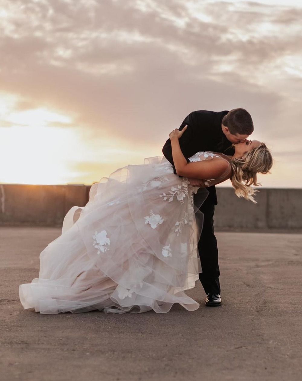 Couple in wedding attire sharing a dip kiss at sunset on an open pavement.