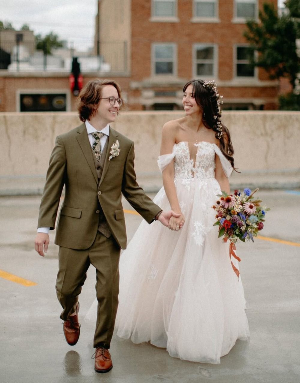 Two brides holding hands, smiling in wedding attire, walking outdoors with a colorful bouquet