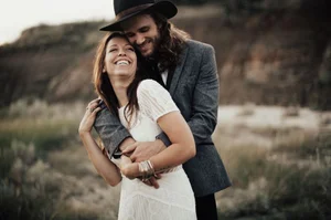 Couple embracing outdoors, smiling in a grassy, blurred landscape, the man wearing a hat and blazer.