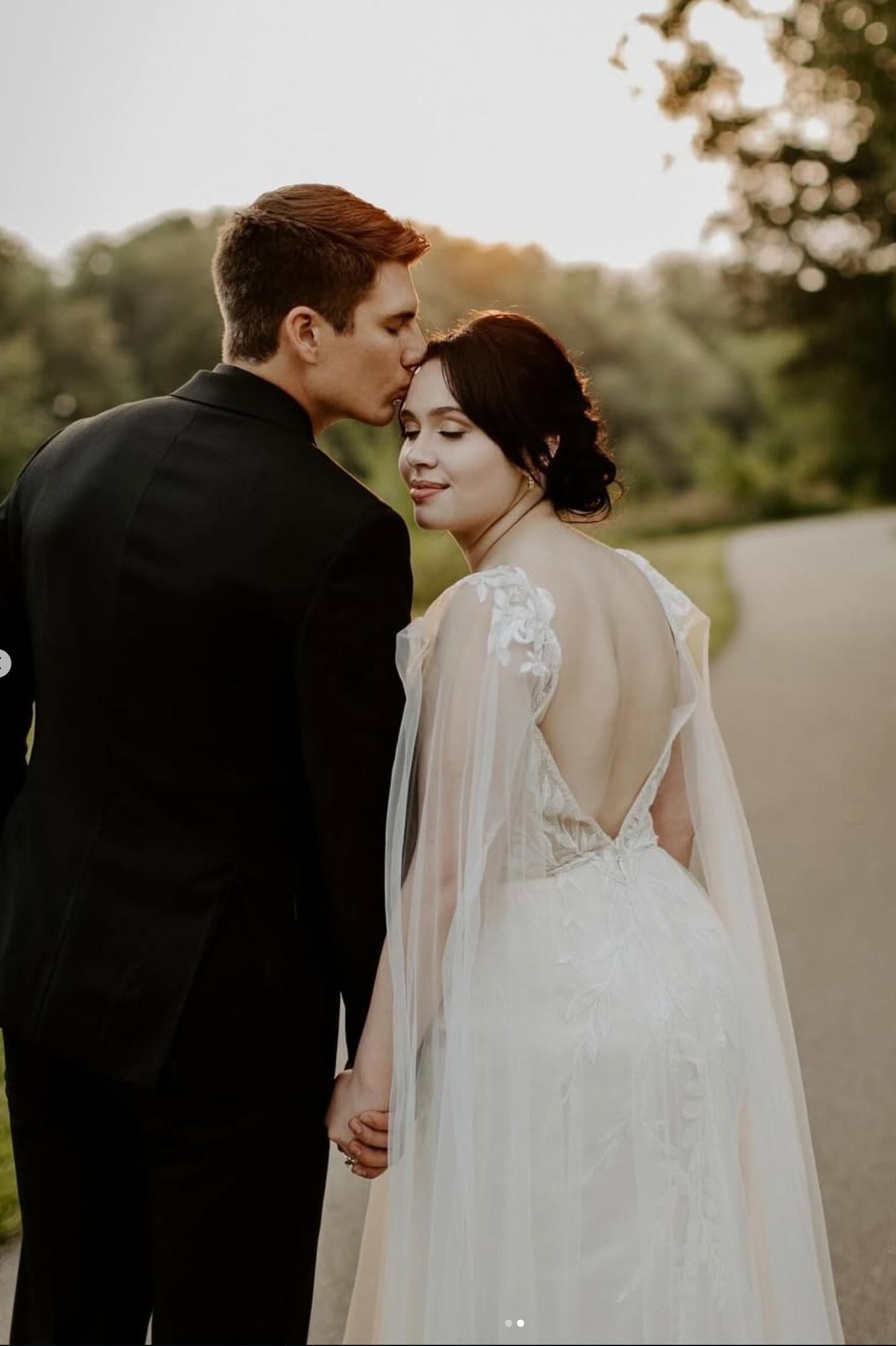 Couple in formal attire outdoors, with the man kissing the woman on the forehead, back view of her lace dress.