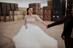 Bride in a white wedding dress holding hands in an industrial outdoor setting, smiling amid stacked crates