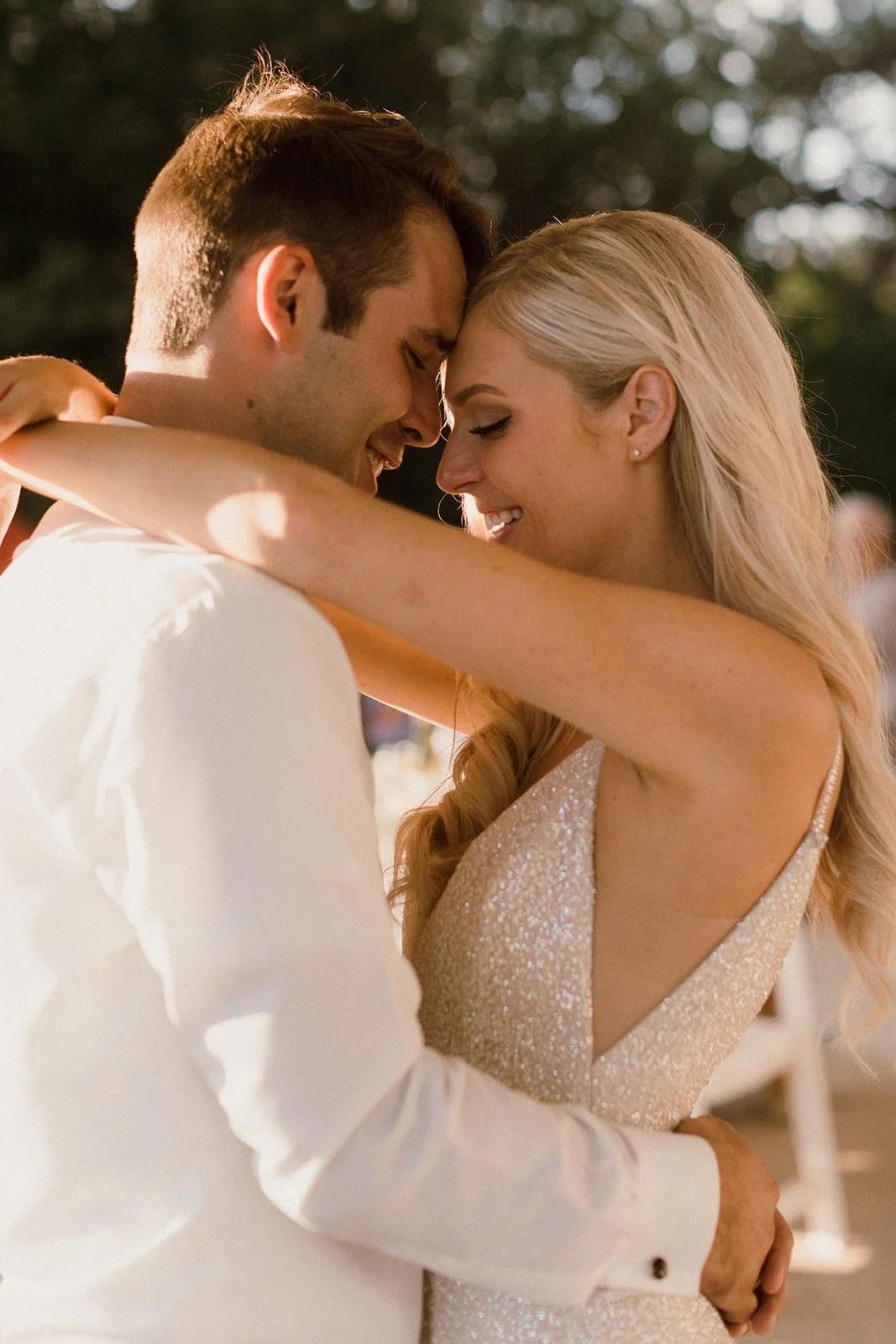 A couple in formal attire slow dancing closely outdoors at a sunlit wedding.