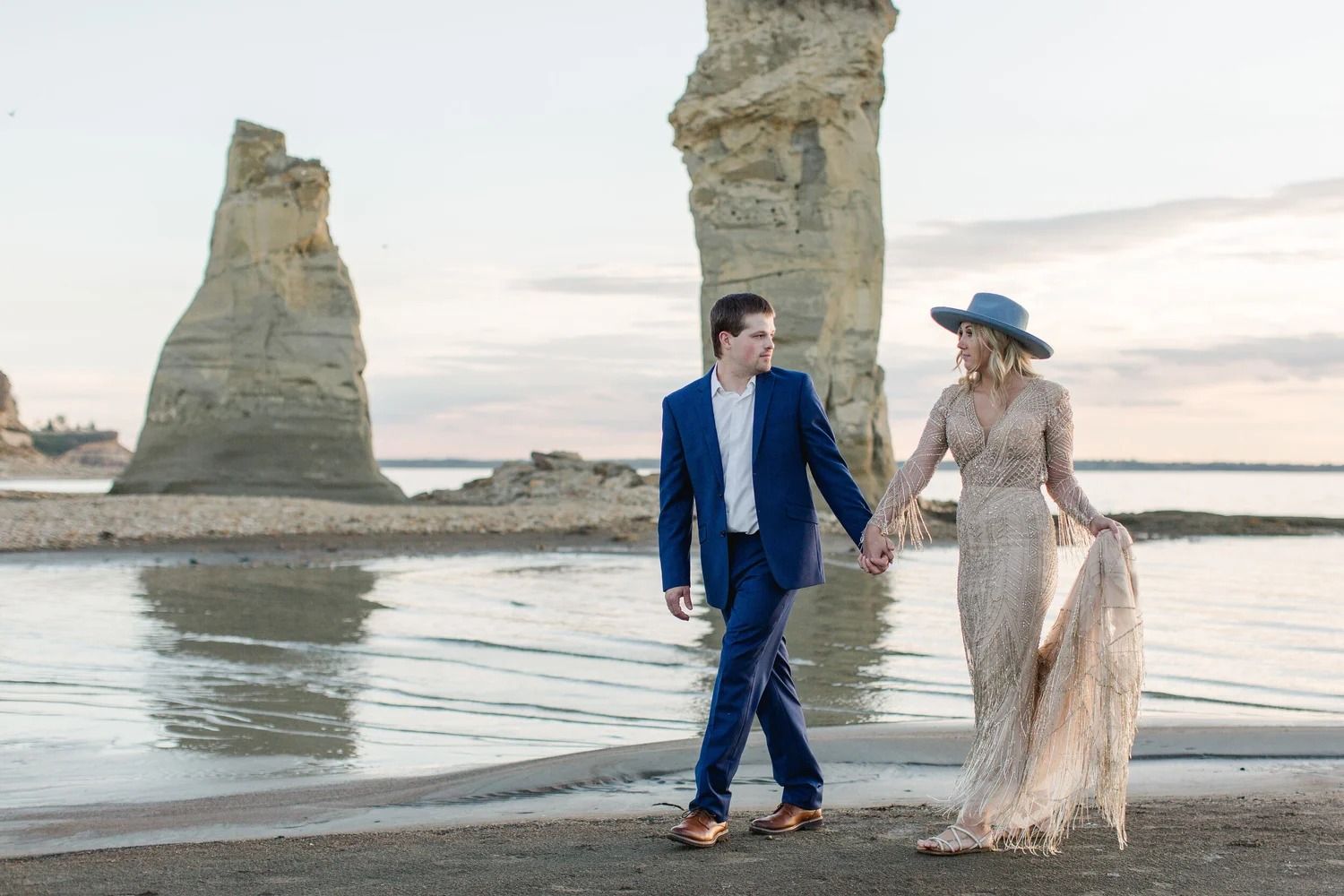 Couple walking hand in hand on a beach beside tall rock formations at sunset
