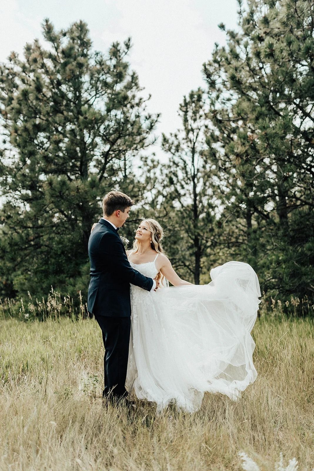 Couple in wedding attire standing in a grassy field with tall pine trees behind them