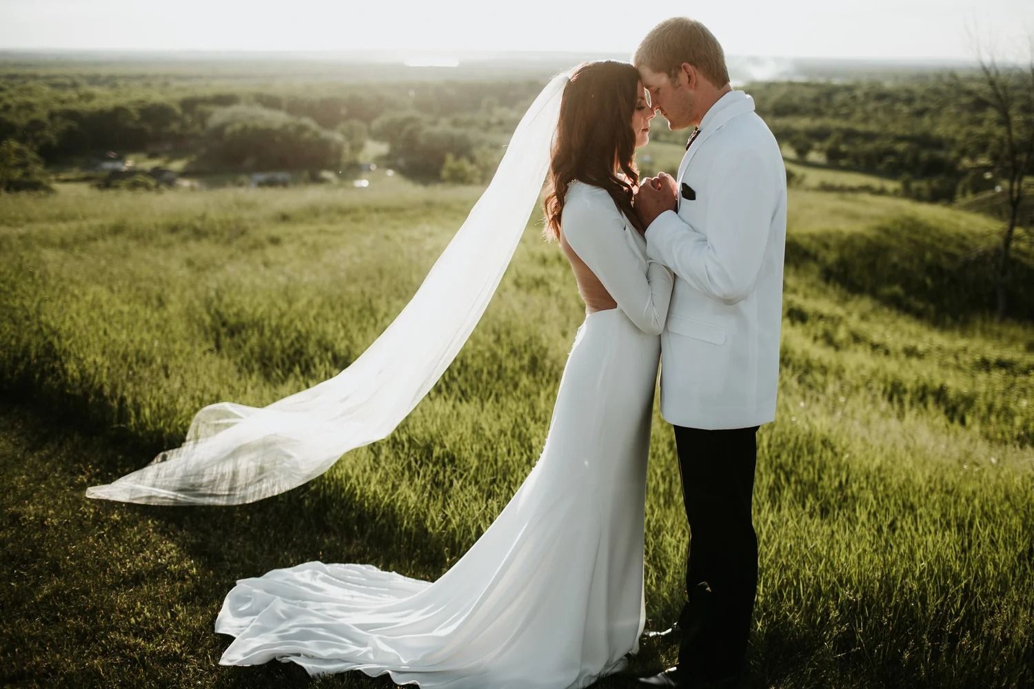 Bride and groom in white attire embracing in a grassy field at sunset