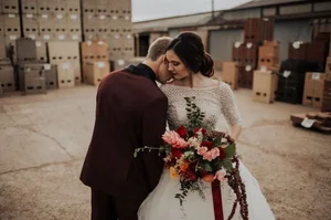 Wedding couple embracing in an industrial courtyard, bride holding a colorful bouquet and wearing a lace gown.