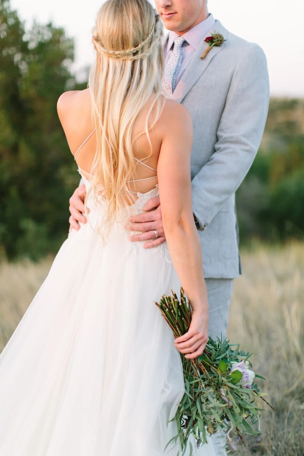 Bride and groom embracing outdoors, with the bride holding a bouquet and wearing a backless white gown