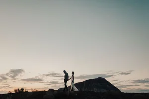 Silhouetted hikers on a ridge at sunset beside a mountain peak under a cloudy sky