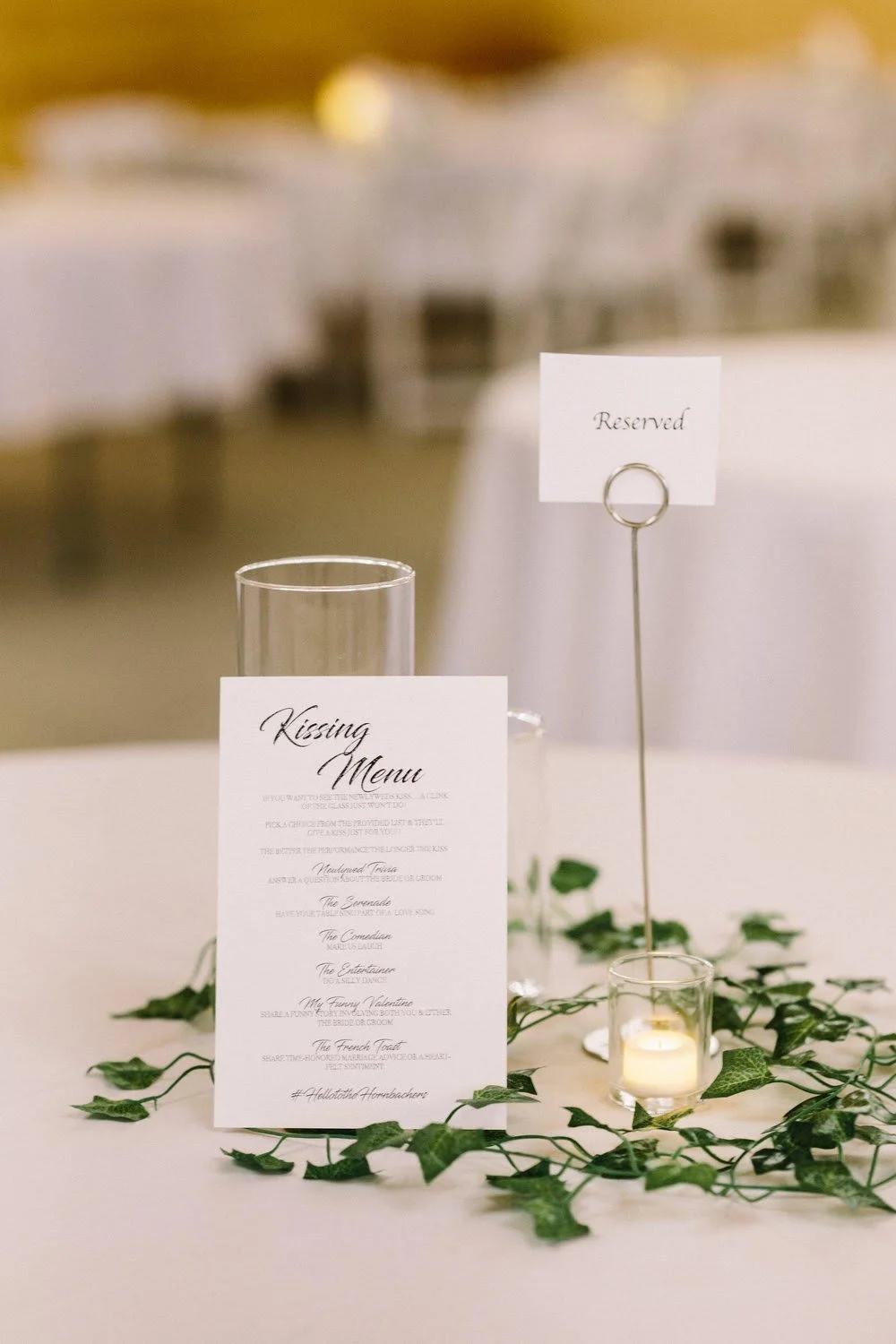 Wedding reception table centerpiece with menu card, table number, candle, and greenery on a white tablecloth
