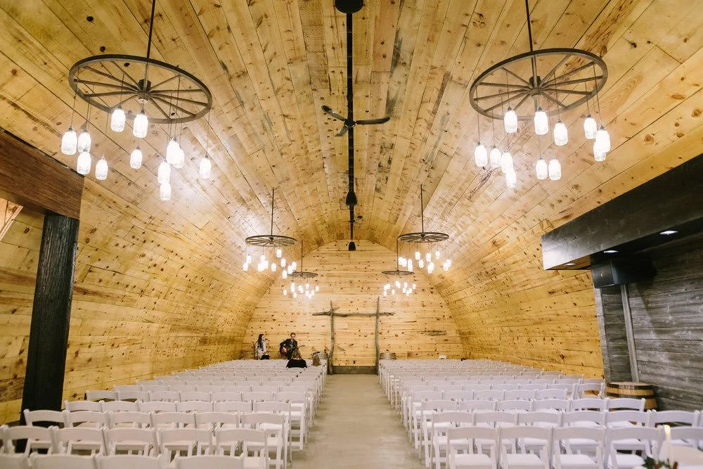 Empty rustic wooden event hall with rows of white chairs and chandeliers under a curved ceiling