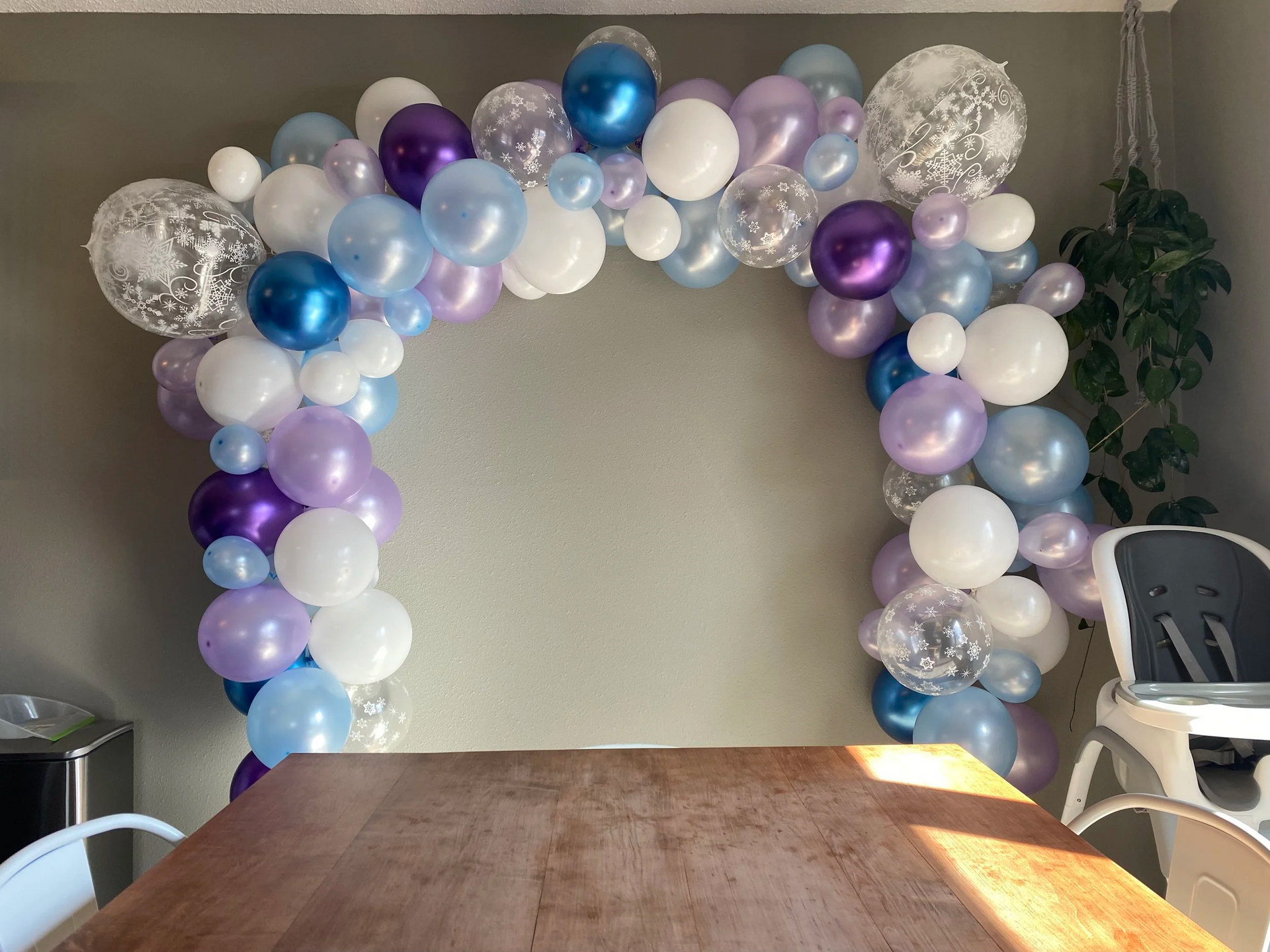 Balloon arch in white, silver, blue, purple, and lavender behind a wooden table indoors