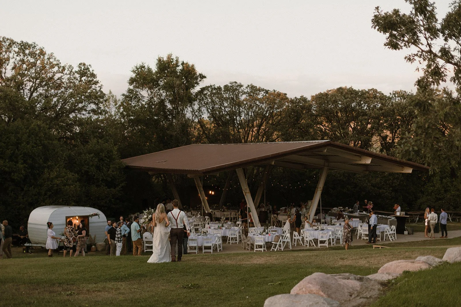 Outdoor wedding reception under a pavilion at dusk, with guests seated on a lawn near trees.