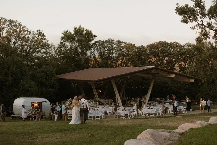 Outdoor wedding reception with guests under a modern pavilion at dusk, white chairs and tables on a lawn.
