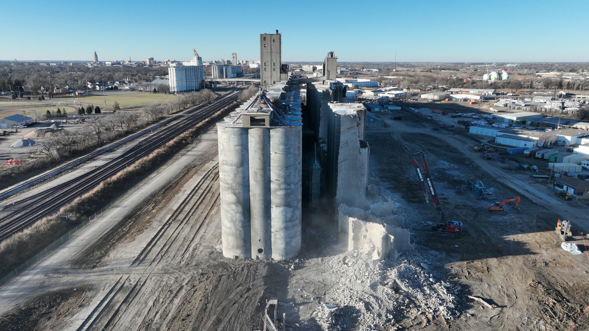 CL Construction demolition crew dismantling the historic Hansen-Mueller grain elevator complex at 3001 Cornhusker Highway in Lincoln, Nebraska. Heavy machinery, including a high-reach excavator with demolition claw, tears down sections of the massive concrete silos and storage structures on the 17.5-acre site south of Cornhusker Highway near 33rd Street, as part of a year-long project to clear the dilapidated landmark for future redevelopment