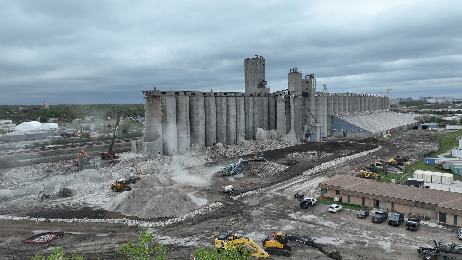 A series of concrete grain silos under demolition with construction equipment and debris on a cloudy day.