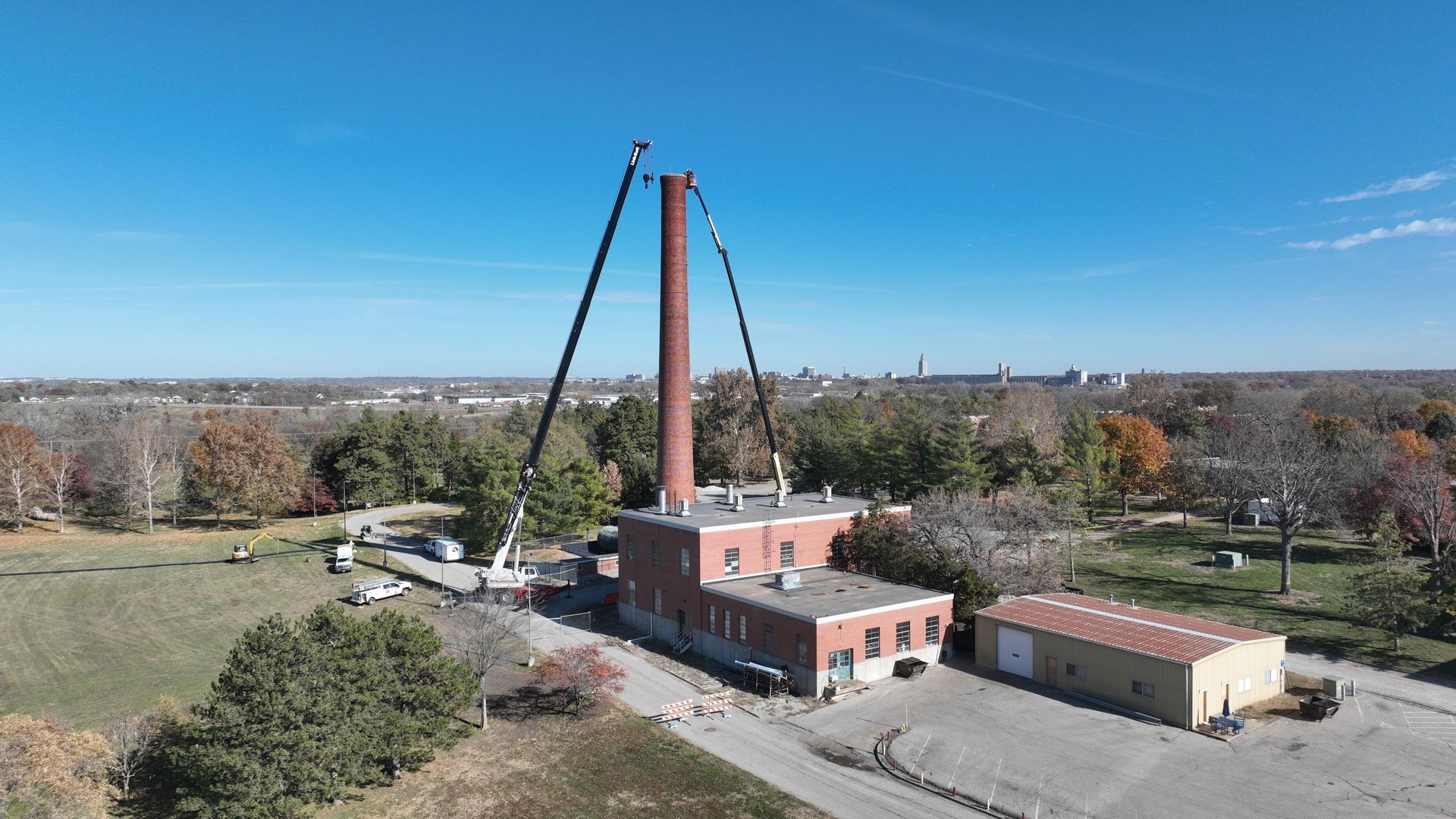 CL Construction crew performing hand demolition on a tall industrial smokestack at the Lincoln Regional Center site in Lincoln, Nebraska. Workers use manual tools and safety gear to carefully dismantle sections of the 143-foot chimney structure amid rubble and heavy equipment on the ground